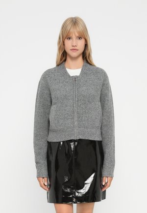 Femme blonde portant un pull en maille gris zippé et une jupe en vinyle noire brillante, se tenant devant un fond blanc uni.