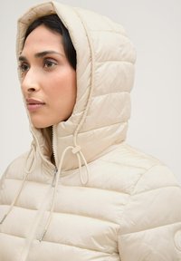 Veste rembourrée beige avec capuche, fermeture éclair et cordons de serrage. Le tissu semble lisse avec des détails de matelassage horizontal.