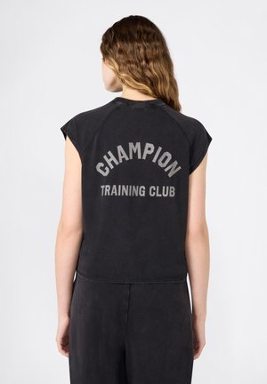 Mujer con cabello ondulado que lleva una camiseta sin mangas negra con el texto "CHAMPION TRAINING CLUB" en la espalda, sobre un fondo liso.