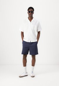White short-sleeve button-up shirt, dark navy shorts, white sneakers with light brown accents, and black sunglasses. Clean, casual look.
