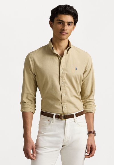 Chemise bouton fermée marron à manches longues, avec un col et un petit logo bleu sur la poitrine, associée à un jean blanc et une ceinture marron.