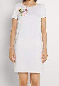 Biała sukienka z krótkim rękawem i okrągłym dekoltem, zdobiona kolorowym haftem kwiatowym na piersi, wykonana z miękkiego bawełnianego materiału.