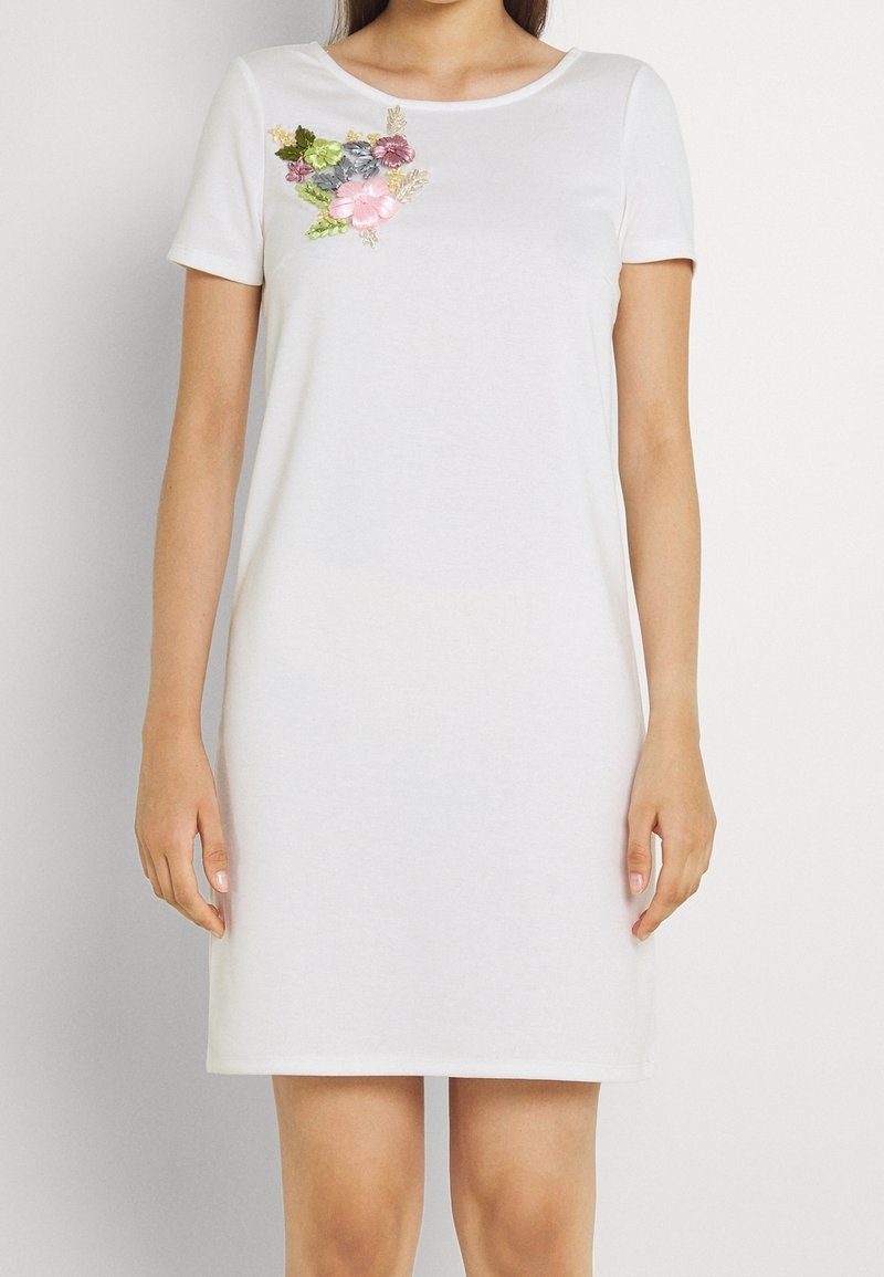 Biała sukienka z krótkim rękawem i okrągłym dekoltem, zdobiona kolorowym haftem kwiatowym na piersi, wykonana z miękkiego bawełnianego materiału.