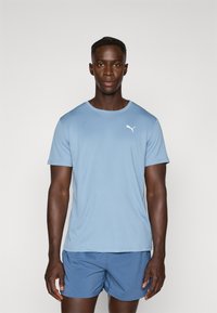 T-shirt bleu clair à manches courtes en tissu doux, arborant un logo Puma blanc sur la poitrine gauche. Porté avec un short de sport bleu.