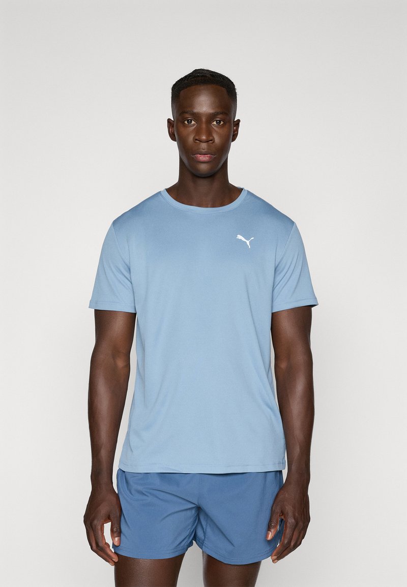 T-shirt bleu clair à manches courtes en tissu doux, arborant un logo Puma blanc sur la poitrine gauche. Porté avec un short de sport bleu.