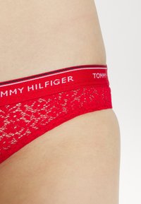 Perizoma in pizzo rosso con un elastico a righe rosse e bianche con il logo "TOMMY HILFIGER". Il materiale è delicato e presenta un motivo floreale.