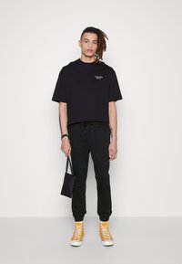 Schwarzes Baumwoll-T-Shirt mit kurzen Ärmeln und einem Logo, kombiniert mit schwarzen Jogginghosen mit Kordelzug und gelben High-Top-Sneakern, während eine kleine schwarze Tasche gehalten wird.
