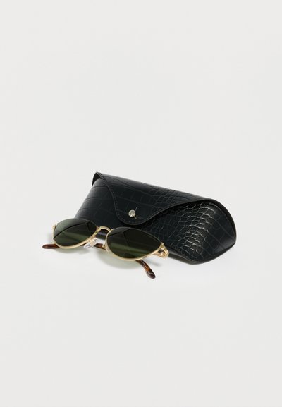 Οβάλ γυαλιά ηλίου με χρυσό σκελετό και σκουρόπρασινους φακούς δίπλα σε μαύρη υφασμάτινη θήκη πάνω σε λευκή επιφάνεια.