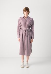 Abito a righe bordeaux e bianco con vita accentuata da una cintura, maniche lunghe, chiusura a bottoni e design di lunghezza media. Abbinato a sneakers bianche.