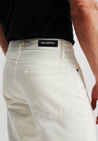 Jeans de mezclilla blanca con corte recto, que cuentan con un diseño de bolsillo trasero, tela texturizada y una etiqueta de logo negra en la cinturilla.