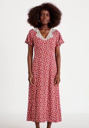 Vrouw met natuurlijk krullend haar die een rode midi-jurk met bloemenprint draagt, korte mouwen en een V-hals met kantafwerking, staand voor een effen achtergrond.