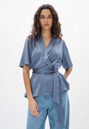 Top en bleu clair à enrouler, fabriqué dans un tissu texturé, avec des manches courtes, une taille nouée et un décolleté en V. Associé à un jean en denim.