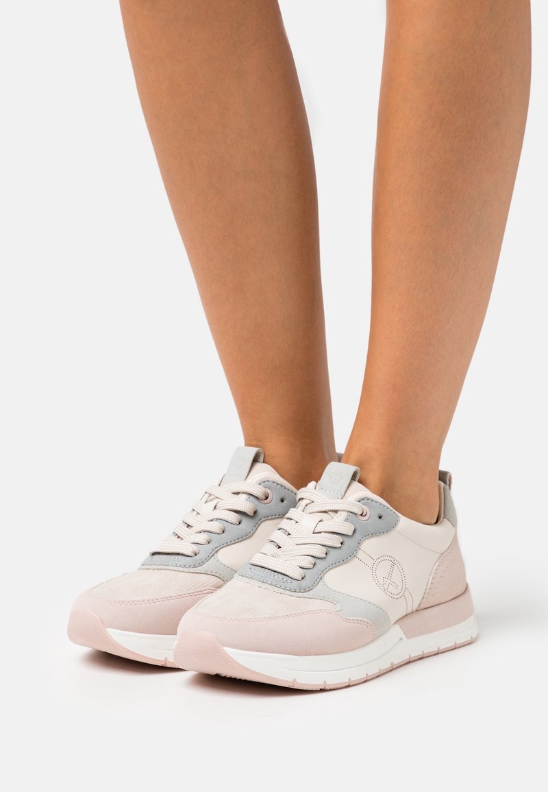 Des baskets à lacets rose pâle et gris portées pieds nus, présentées sur un fond blanc uni.