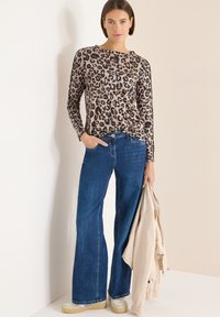 Beżowa i czarna bluzka w panterkę z długim rękawem, zestawiona z niebieskimi dżinsami o szerokich nogawkach. Modelka trzyma lekką beżową kurtkę.