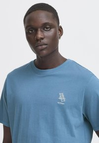 Blå bomulds t-shirt med rund halsudskæring, korte ærmer, og et lille hvidt broderet stol-design med ordet "RETREAT."