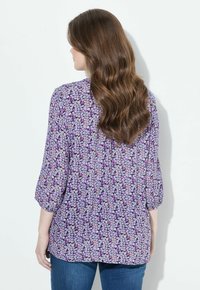 Lila Bluse mit dreiviertel Ärmeln, aus einem leichten, strukturierten Stoff und mit kleinen, mehrfarbigen Blumenmustern.