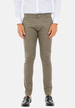 Uomo che indossa pantaloni marroni slim fit a quadri, camicia bianca elegante e mocassini neri in pelle, in piedi contro uno sfondo semplice.