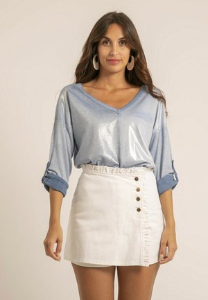 Femme debout, portant un chemisier bleu clair brillant à manches longues avec des poignets retroussés et une jupe blanche taille haute avec des boutons sur un côté.