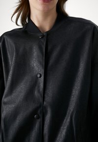 Femme portant une veste noire en similicuir avec un col montant et des fermetures à boutons-pression sur un fond blanc uni.