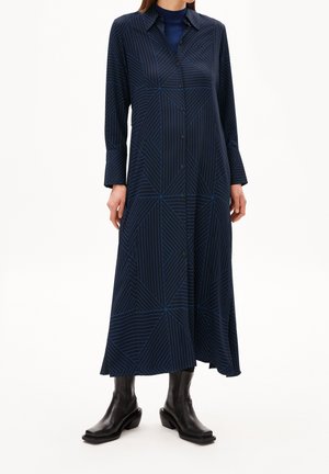 Vrouw in een lange marineblauwe jurk met geometrische blauwe strepen, met knopen aan de voorkant, lange mouwen, gecombineerd met zwarte laarzen tot halverwege het kuit.