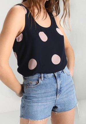 Femme portant un haut sans manches bleu marine avec de gros pois rose pâle et un short en jean bleu clair taille haute avec des ourlets effilochés.