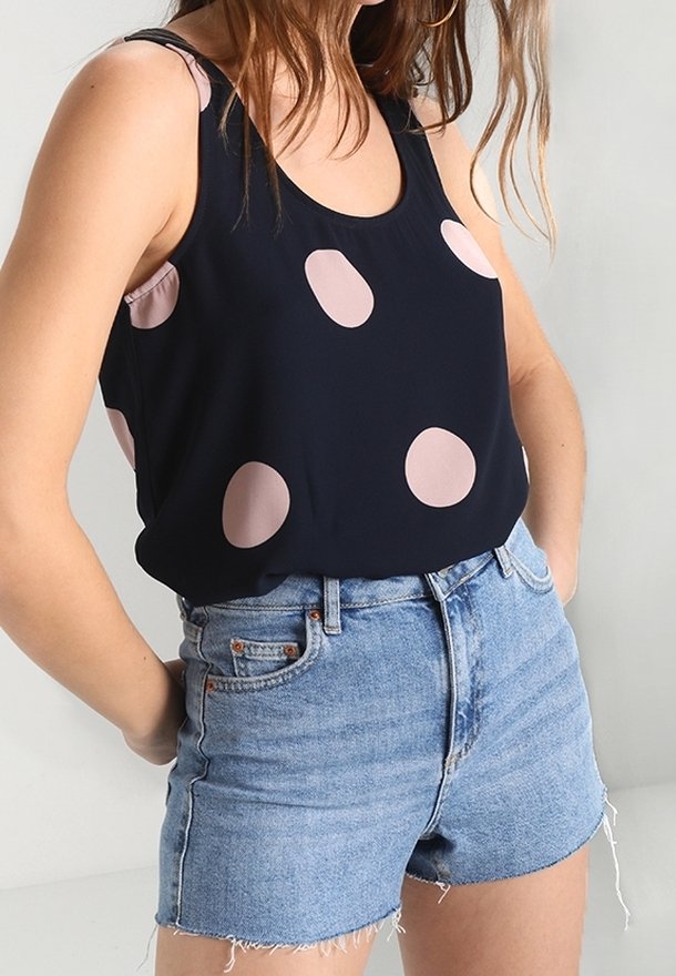 Femme portant un haut sans manches bleu marine avec de gros pois rose pâle et un short en jean bleu clair taille haute avec des ourlets effilochés.