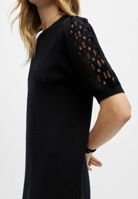Schwarzes Kleid mit kurzen Ärmeln und hexagonalen Ausschneidungen. Weicher Stoff, taillierte Silhouette, Rundhalsausschnitt und seitliche Schlitze.