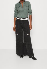 Vihreä nappipaitapusero, jossa on rintataskut, yhdistetty mustiin leveälahkeisiin cargo-housuihin, joissa on vetskari ja valkoinen vyö. Mustat korolliset saappaat.