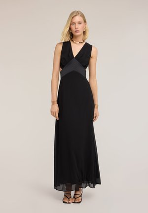 Lyshåret kvinde iført en sort maxikjole uden ærmer med udskæringer ved taljen, kombineret med sorte hæle med stropper og guldsmykker.