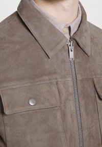 Ruskea mokkatakki, jossa on kauluksellinen kaula-aukko, etuvetoketju ja kaksi rintataskua, joissa on hopeiset nepparit. Pehmeä rakenne ja hienovarainen kiilto.