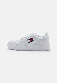Zapatilla blanca con suave cuero sintético, diseño de cordones, perforaciones en la puntera y un parche con el logo en rojo, blanco y azul en el lateral.