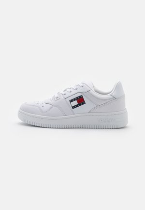 Zapatilla blanca con suave cuero sintético, diseño de cordones, perforaciones en la puntera y un parche con el logo en rojo, blanco y azul en el lateral.