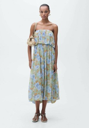 Floral maxi dress in sheer green fabric featuring blue and orange flowers, cinched waist, and adjustable spaghetti straps. Worn with a beige bag.