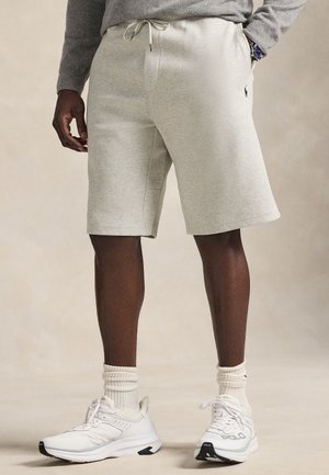 Shorts en coton gris avec une ceinture élastique, des poches latérales et un petit logo sur la jambe gauche, associés à des chaussures de sport blanches et des chaussettes côtelées.