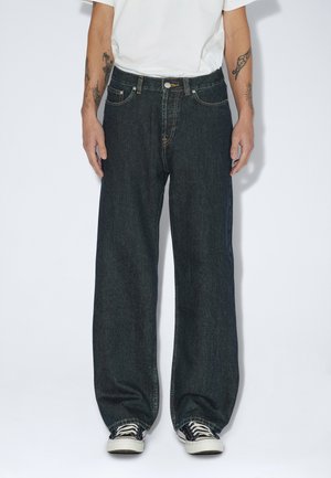 Person iført løstsiddende mørke denimjeans, hvide sneakers, en hvid skjorte synlig fra taljen og ned, med tatoverede arme ned langs siden.