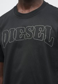 Detailný záber na čierne tričko s nápisom "DIESEL" vyšité veľkými, obrysnými písmenami cez hruď.