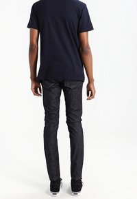 Persona de espaldas, vistiendo una camiseta oscura y unos jeans oscuros, de pie contra un fondo blanco, con zapatos informales visibles.