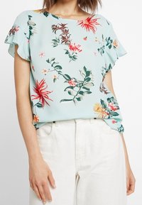 Květinová blůza s krátkým rukávem ze světle modré látky, s živými vzory červených, žlutých a zelených květin. Lehké provedení a volný střih.