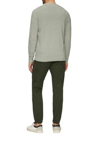 Homme portant un pull gris clair, un pantalon cargo vert foncé avec des poches à rabat, et des baskets blanches avec des accents bleu marine, se tenant devant un fond blanc.
