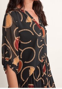 Svart klänning med V-urringning, korta ärmar, med ett mönster av cirklar och snorlika mönster i rost- och tanfärger. Mjukt, lätt tyg.