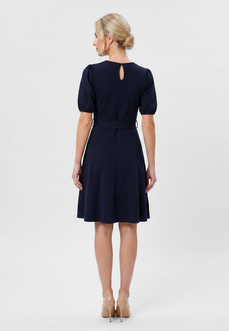 Femme blonde avec les cheveux en chignon portant une robe bleu marine longueur genou avec des manches ballon et des chaussures à talons beiges, vue de dos.