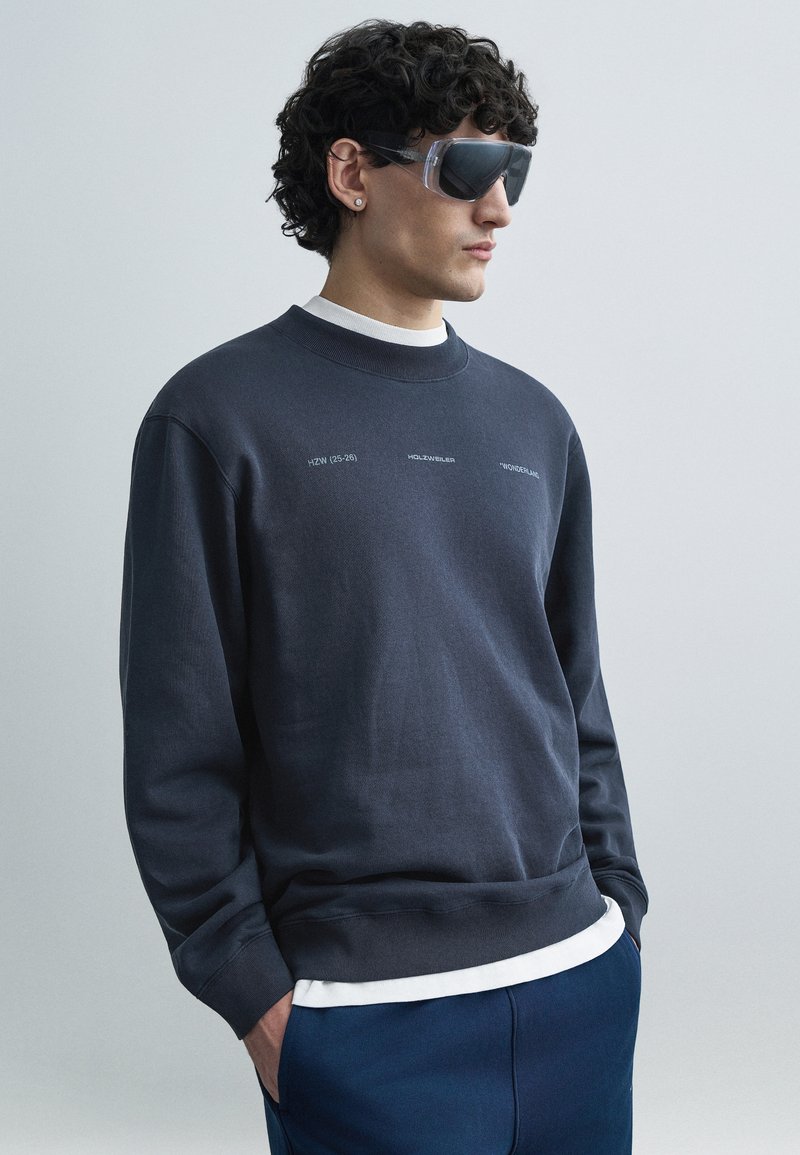 Mornarsko modra jopa z okroglim izrezom, okrašena z belim tiskanim besedilom. Model nosi prozorna sončna očala; modre hlače z žepi. Gladek tekstura.