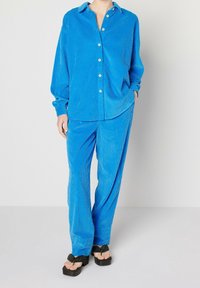 Sininen corduroy-paita, jossa on napitettava paita ja yhteensopivat housut. Ominaisuuksina rento malli, pitkät hihat ja sivutaskut. Litteät sandaalit.