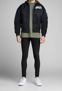 Chaqueta bomber negra con puños acanalados, bolsillos con cremallera y texto gráfico blanco en el pecho, combinada con una camisa verde oliva y leggings negros.