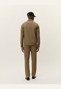 Completo giacca e pantaloni marrone, realizzato in tessuto liscio. La giacca presenta un colletto alto e dettagli di cucitura visibili. Un cappellino nero completa l'outfit.