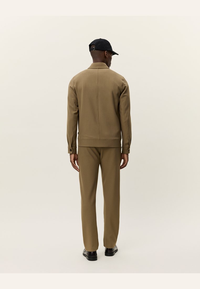 Completo giacca e pantaloni marrone, realizzato in tessuto liscio. La giacca presenta un colletto alto e dettagli di cucitura visibili. Un cappellino nero completa l'outfit.