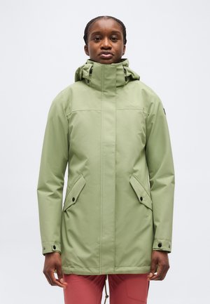 Personne portant une veste de pluie à capuche vert clair avec poches avant et poignets boutonnés, debout devant un fond clair uni.