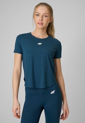 Młoda kobieta z blond kucykiem, ubrana w dopasowaną ciemnoniebieską sportową koszulkę i legginsy z białymi logo 4F na jednolitym szarym tle.