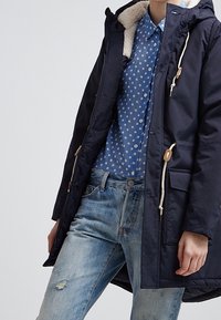 Parka blu navy con cappuccio foderato in pelliccia, dotato di due tasche frontali, bottoni in legno e coulisse. Indossato sopra una camicia blu a pois.