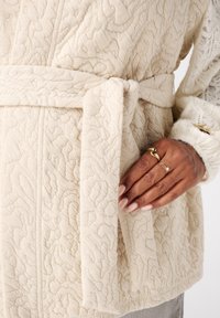 Lichtbeige textuur badjas met een strikceintuur, featuring een verhoogd bloemenpatroon. Hand getoond met delicate ringen en neutrale nagellak.